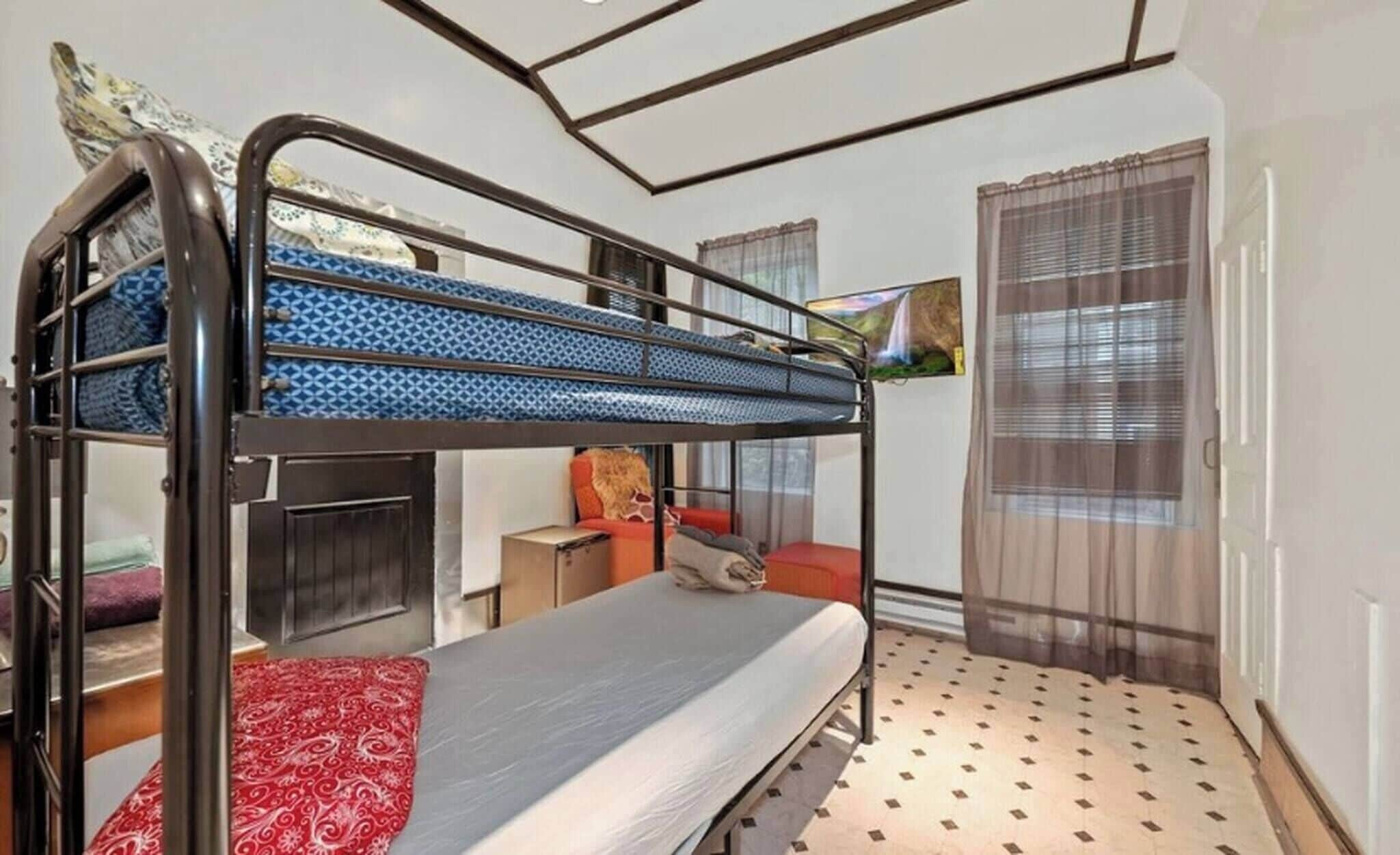 A bunk bed room with a patterned mattress, a red blanket, a small table with a chair, and a painting on the wall.
