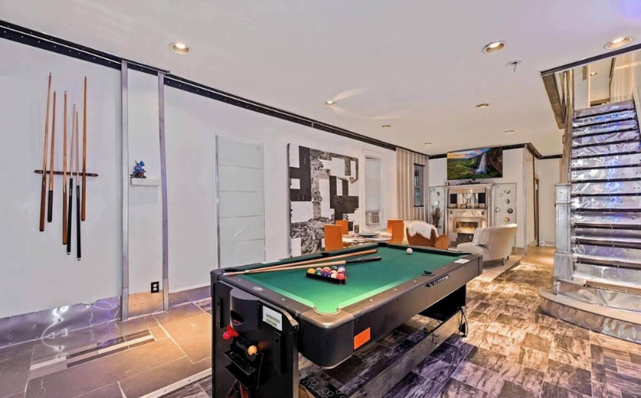 A modern indoor pool table with a green felt surface, a billiards rack, and cue sticks on a wall.