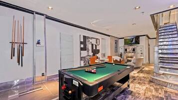 A modern indoor pool table with a green felt surface, a billiards rack, and cue sticks on a wall.