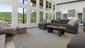 Uma sala de estar espaçosa com grandes janelas, um sofá modular, uma mesa de centro e vista para uma área arborizada.