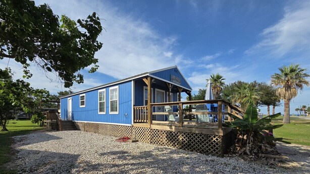 A blue building with a wooden deck, surrounded by gravel and palm trees. - Tiny Home Walking Distance to the Beach (Port O`Connor)