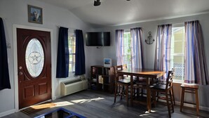 A living room with a wooden table, chairs, a ceiling fan, a TV, and a door with a glass window. - Tiny Home Walking Distance to the Beach (Port O`Connor)