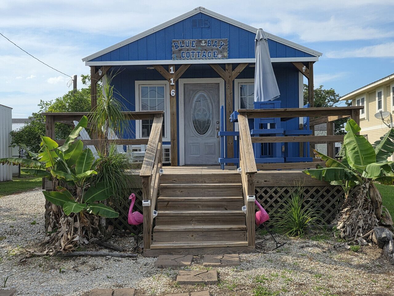 A small, single-story building with a blue exterior, a wooden front porch, and a sign reading 'Blue Bay Cottage.'
