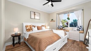 Une chambre à coucher comprenant un lit, une table de chevet, une lampe, une fenêtre avec des rideaux et un ventilateur de plafond.