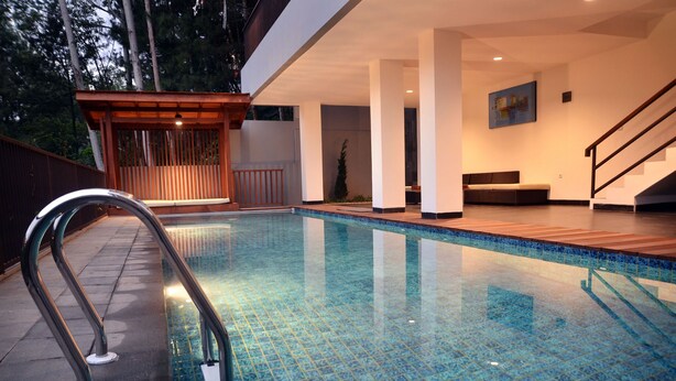 A modern indoor swimming pool with clear blue tiles, a wooden deck, and a staircase leading to an upper level. - Cempaka 2 Villa Dago Private Pool (Bandung)