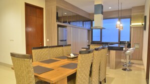A modern dining area with a wooden table, woven chairs, a kitchen in the background, and a large window. - Cempaka 2 Villa Dago Private Pool (Bandung)