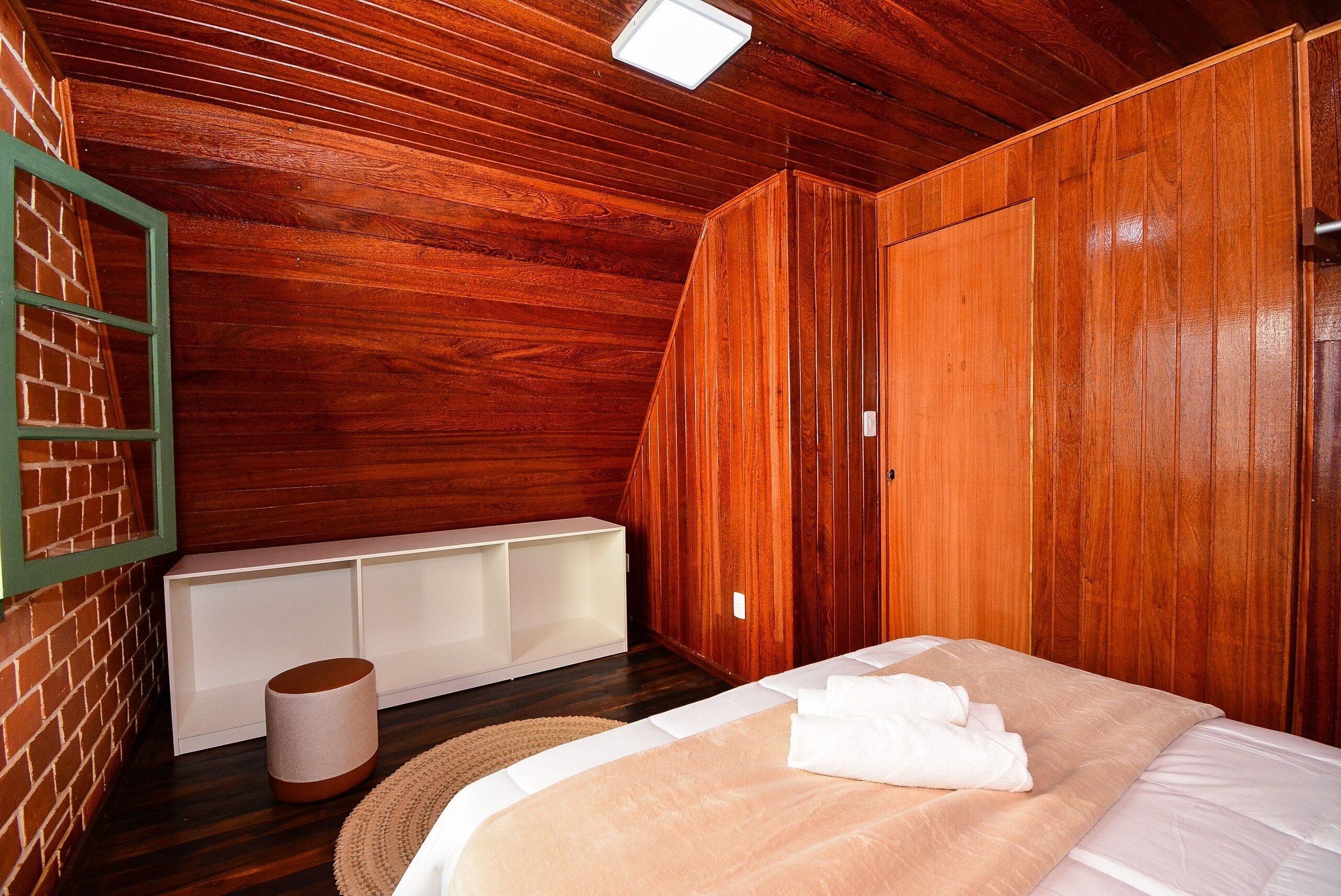 Quarto com painéis de madeira, uma cama, um banco branco e um objeto cilíndrico no chão.