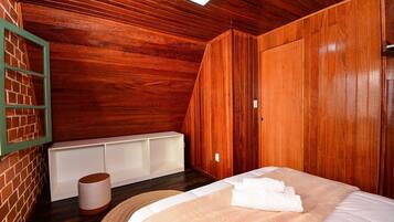 Quarto com painéis de madeira, uma cama, um banco branco e um objeto cilíndrico no chão.