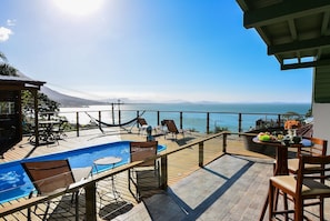 A pool area with a view of the sea and mountains, wooden deck, and outdoor seating.