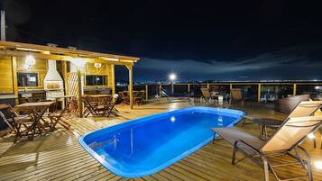 A wooden deck with a swimming pool, lounge chairs, and a barbecue area.