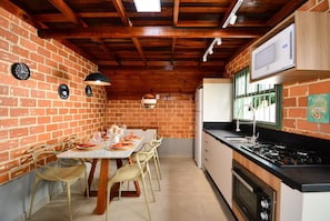 A kitchen with a brick wall, wooden ceiling beams, a dining table with chairs, a refrigerator, a microwave, and a stove.