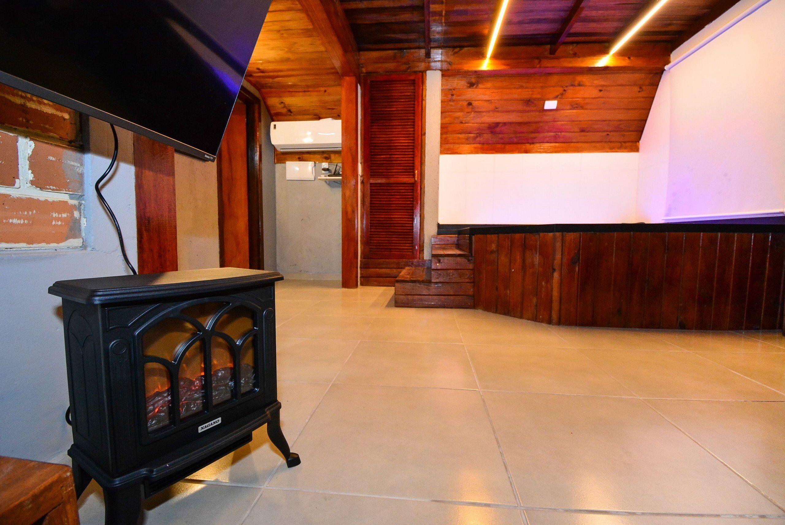 A room with a black stove, wooden walls, a tiled floor, and a staircase leading to an upper level.