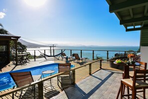 Zona de piscina con vistas al mar y a las montañas, terraza de madera y mobiliario exterior.