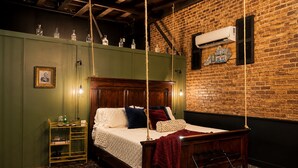 A bedroom with a wooden bed, a green wall, a brick wall, a hanging light fixture, and a small shelf with bottles.