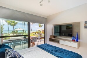 A bedroom with a large bed, a TV mounted on the wall, and a sliding glass door leading to an outdoor area with a pool and palm trees. - Porto Beach Residence (Ipojuca)