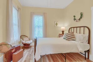 Ein Schlafzimmer mit einem Bett, einem Sessel, einem Nachttisch, einer Lampe, einem Globus und einem Fenster mit Vorhängen.