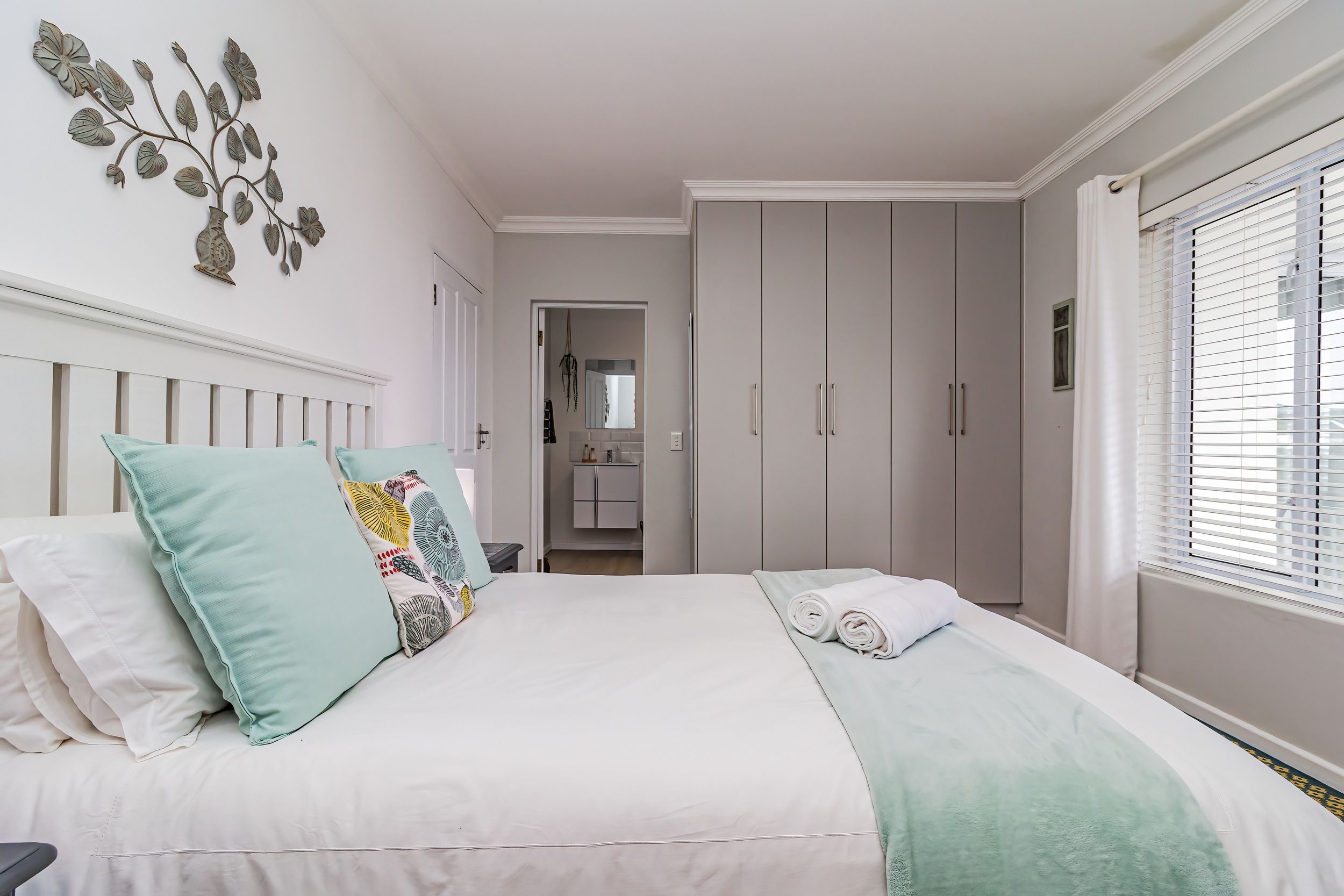 A bedroom with a bed, pillows, a bedside table, a wardrobe, and a window with blinds.