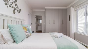 A bedroom with a bed, pillows, a bedside table, a wardrobe, and a window with blinds.
