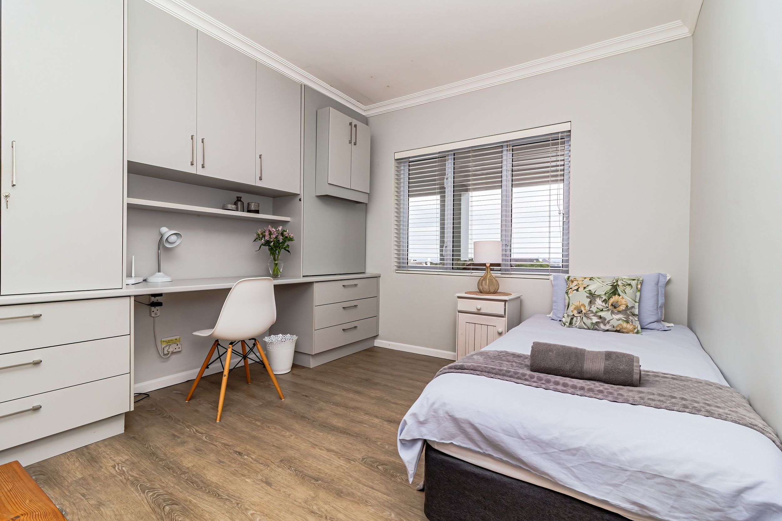 A bedroom with a bed, a desk, a chair, a window with blinds, and a nightstand.