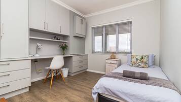 A bedroom with a bed, a desk, a chair, a window with blinds, and a nightstand.