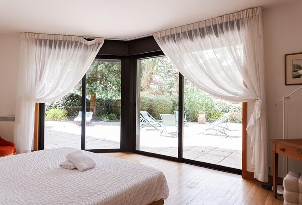 Ein Schlafzimmer mit einem großen Bett, weißen Vorhängen und Blick auf eine Terrasse mit Stühlen und einem Tisch.