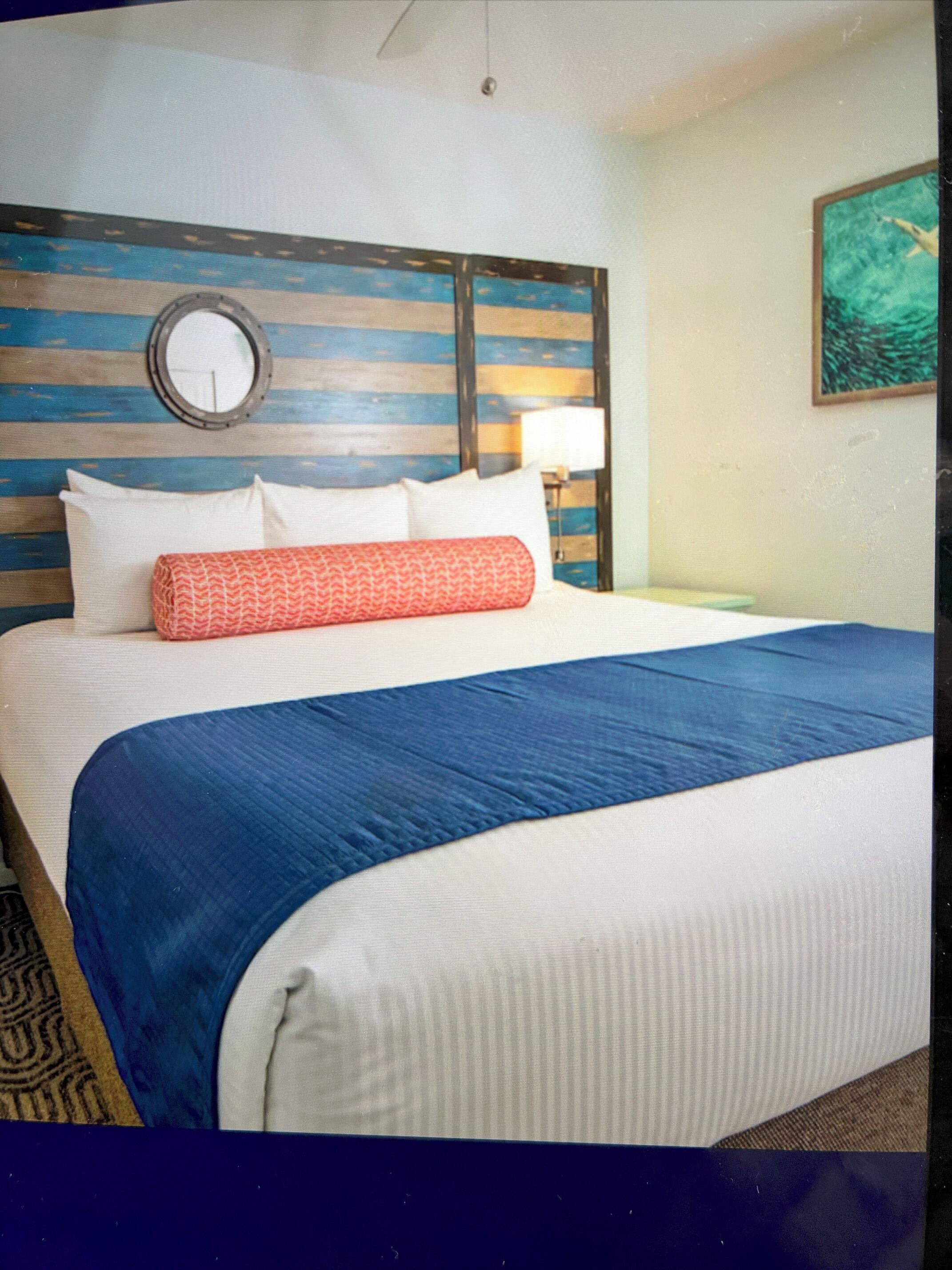 A hotel room with a bed, a headboard with horizontal stripes, a round mirror, a nightstand with a lamp, and a painting on the wall.
