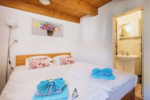 Ein Schlafzimmer mit Holzbalkendecke, einem Bett mit blumigen Kissen, einer Nachttischlampe und einem durchgegangenen Tür erkennbaren Badezimmer.