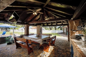 Eine Terrassenanlage im Freien mit Steinboden, Holzmöbeln und einem überdachten Bereich mit Deckenventilator.