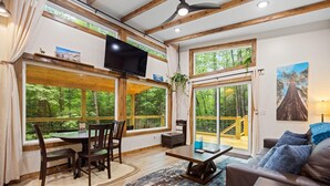 A spacious living room with a large window, a flat-screen TV, a dining area with chairs, a sofa, and a coffee table.