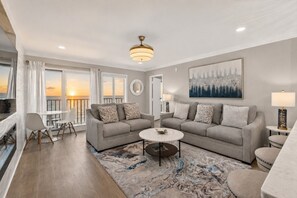 A modern living room with a sofa set, a coffee table, and a view of the sunset through large windows. - Beachfront Condo dr 1402 (Virginia Beach)
