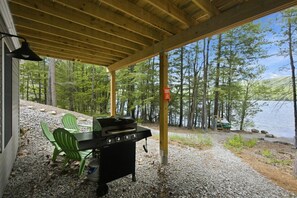 Een overdekt terras met een barbecue, stoelen en uitzicht op een meer en bos.