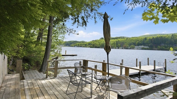 Une terrasse en bois avec des chaises et une table en métal, donnant sur un lac où se trouve un quai.