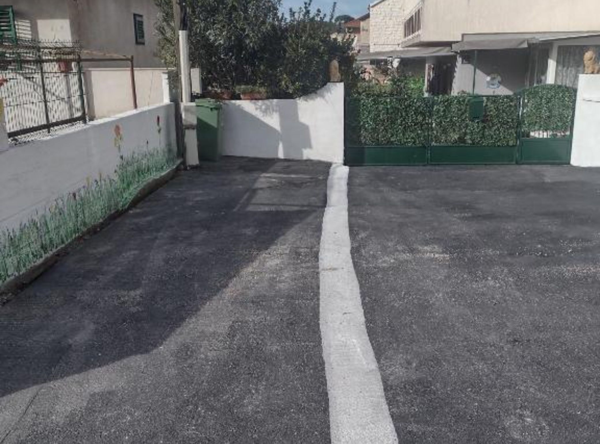 Un patio pavimentado con una línea blanca delimitando, una verja verde y un muro blanco con seto.