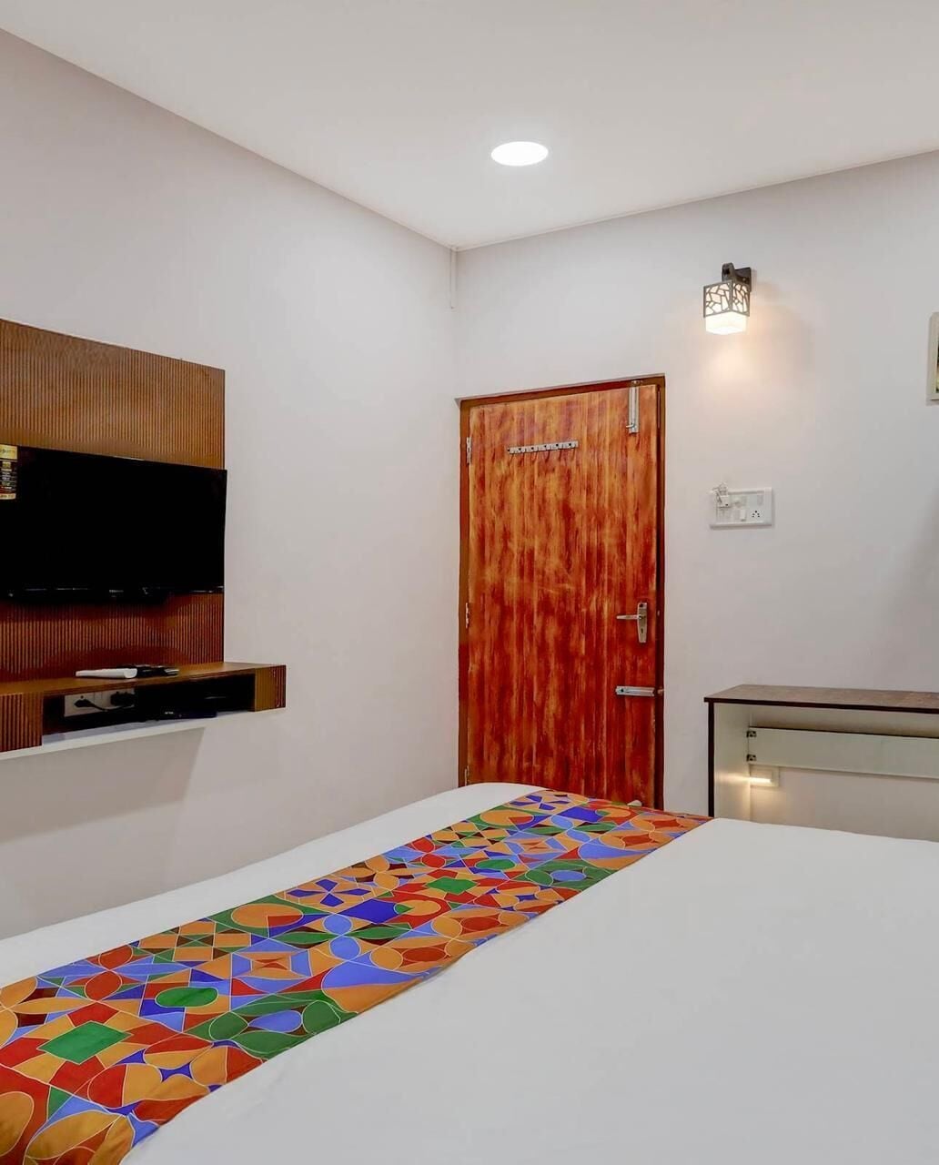 A bedroom with a bed, a television mounted on a wooden panel, a wooden door, and a small cabinet.