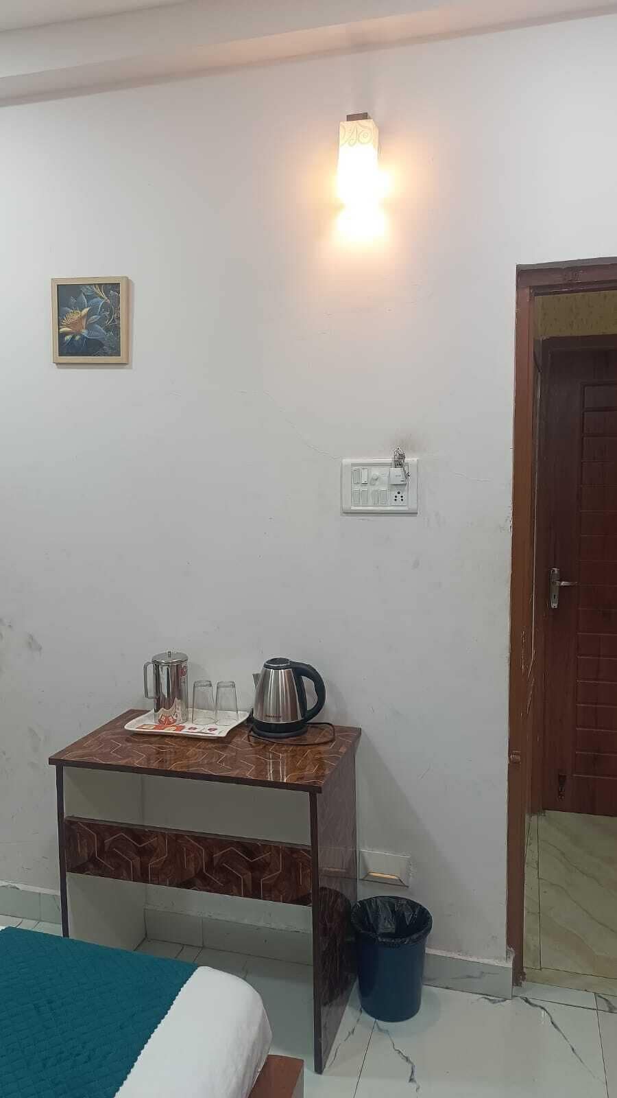 A room with a bed, a small table with a kettle and cups, a wall socket, a trash bin, and a door.