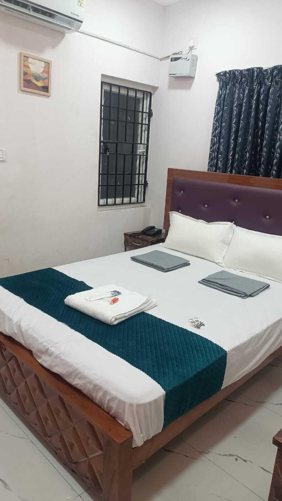 A hotel room with a bed, a window with bars, an air conditioning unit, a bedside table with a phone, and a curtain.