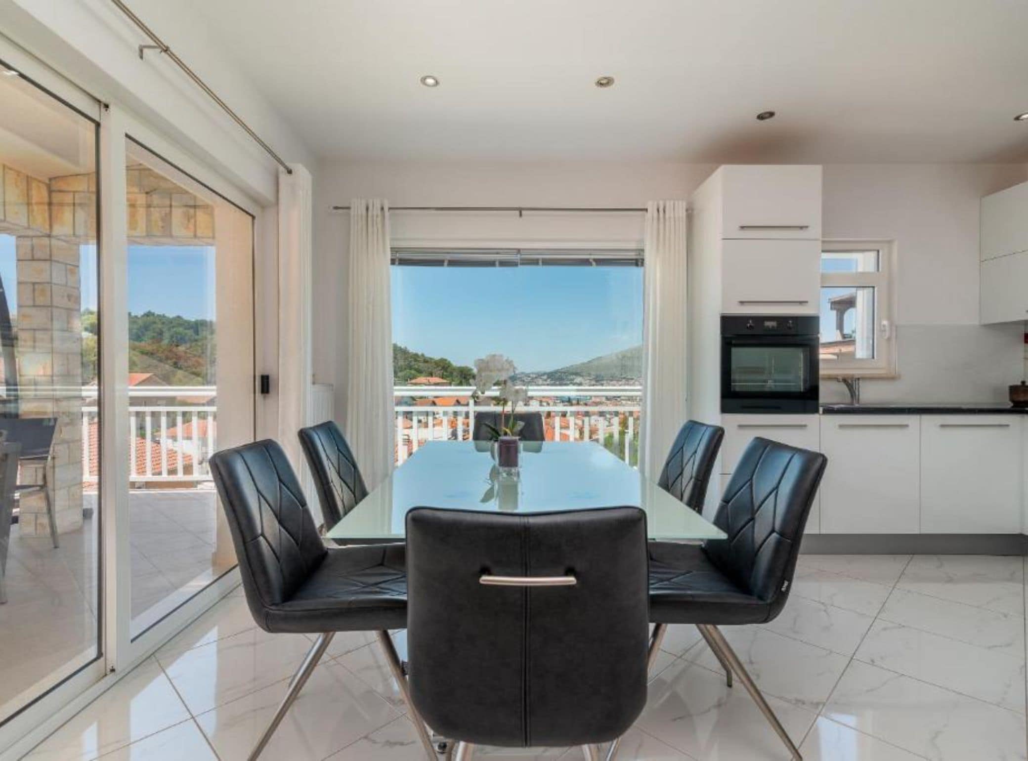 A modern dining area with a glass table and black chairs, a kitchen with white cabinets, and a balcony with a view.