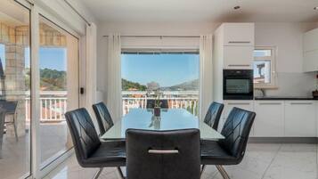A modern dining area with a glass table and black chairs, a kitchen with white cabinets, and a balcony with a view.