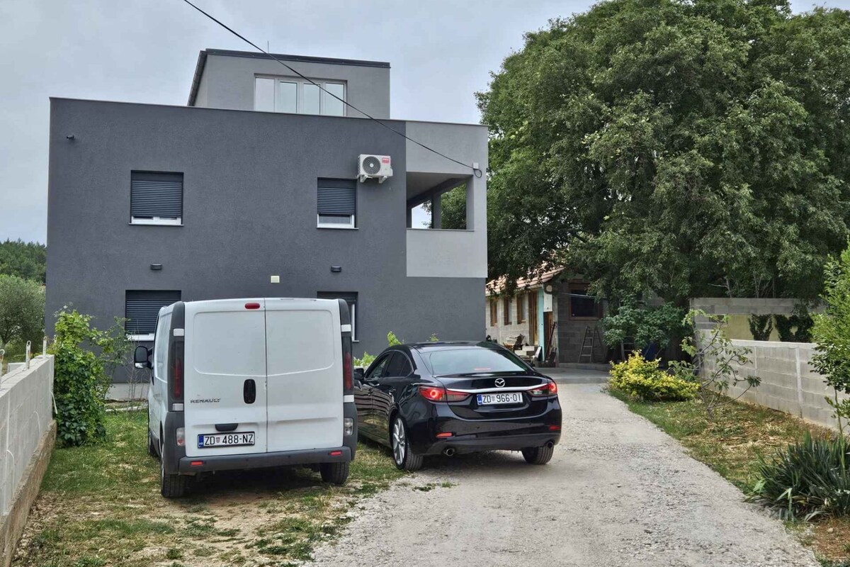 Ein modernes Gebäude mit grauer Fassade, ein weißer Lieferwagen und ein schwarzes Auto auf einem Schotterweg.