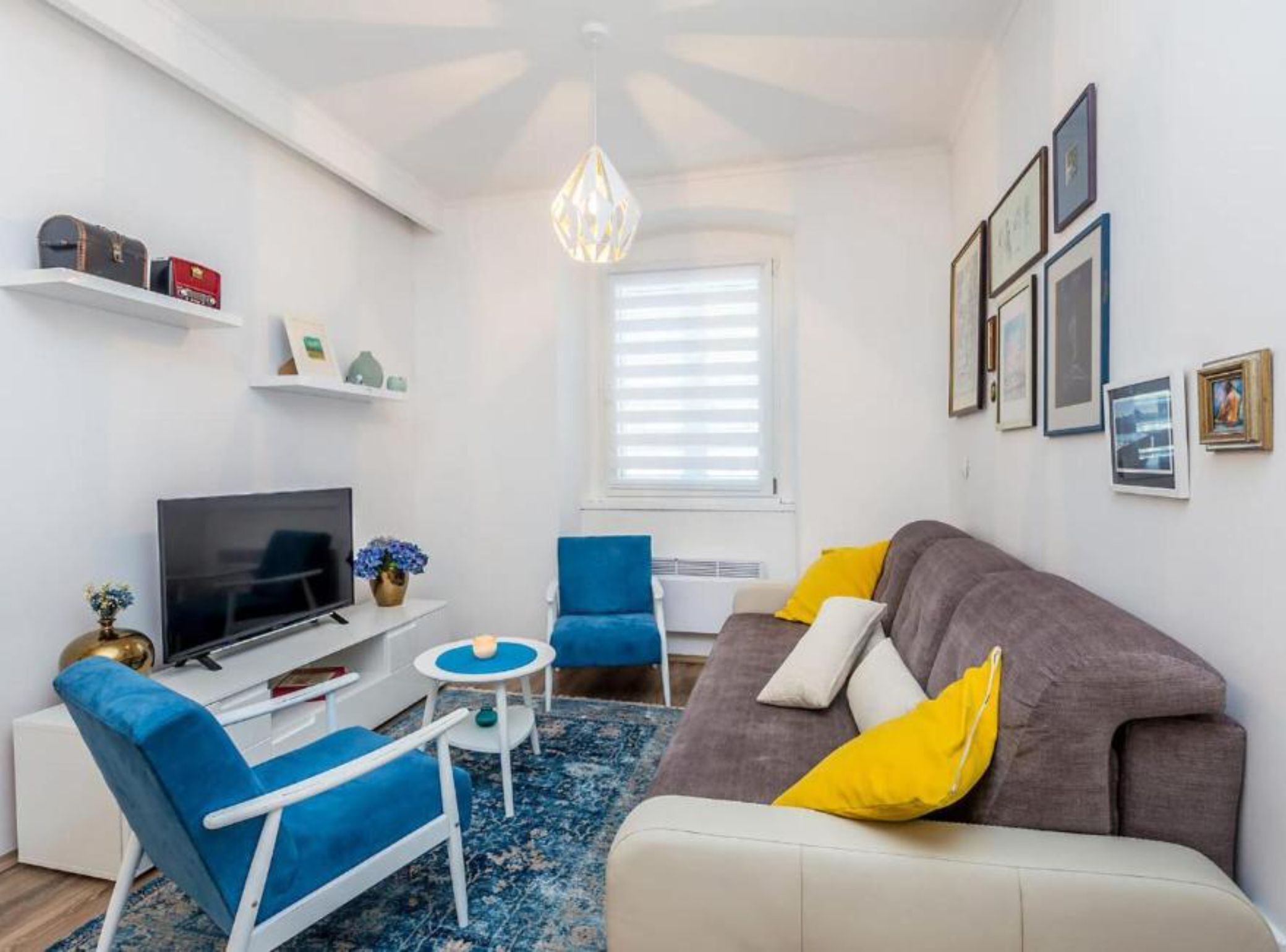 A modern living room with a TV, blue and white furniture, a grey sofa with yellow pillows, and a chandelier.