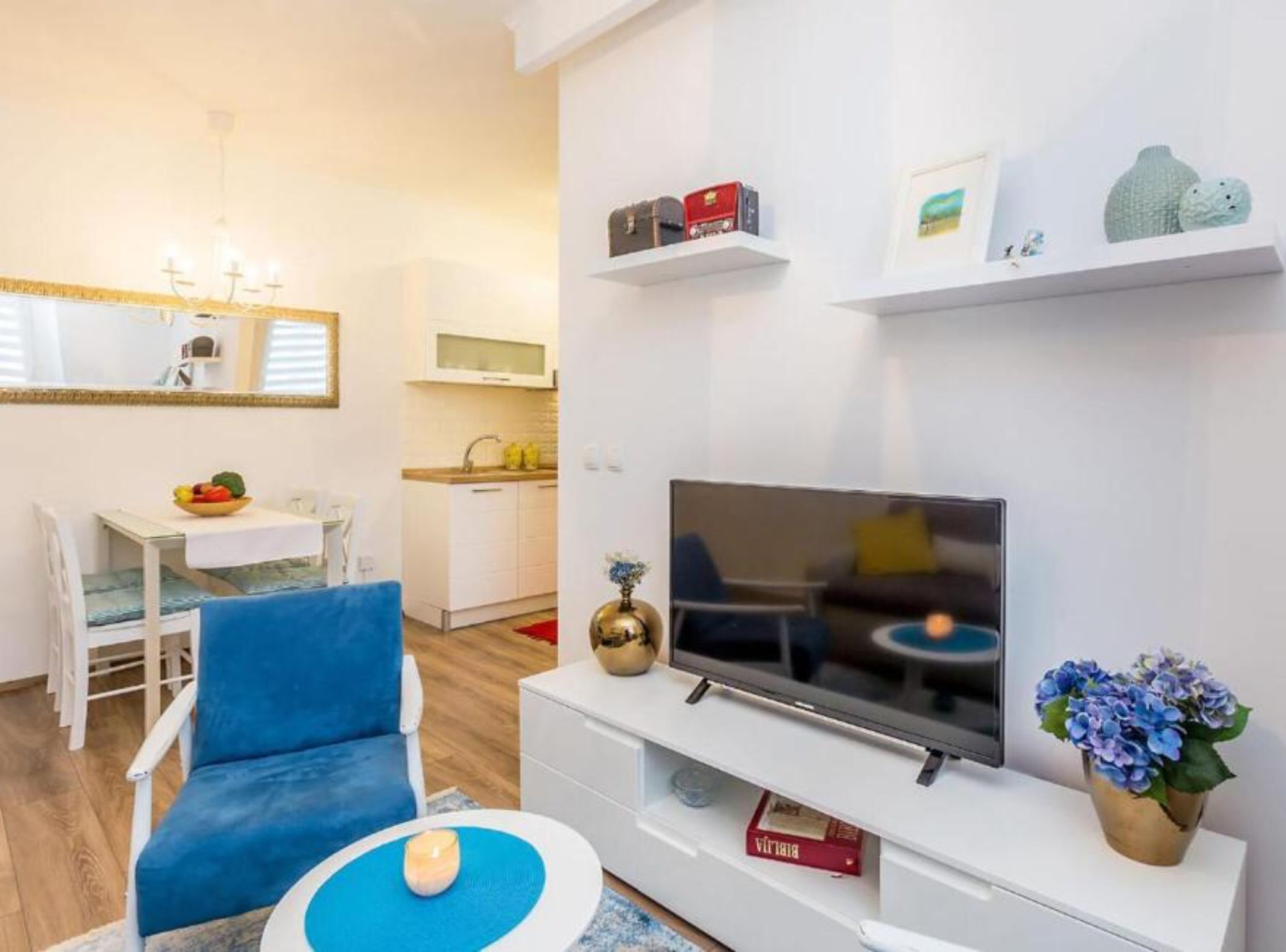 A modern living room with a TV, a blue chair, a small table with a candle, and a kitchen in the background.