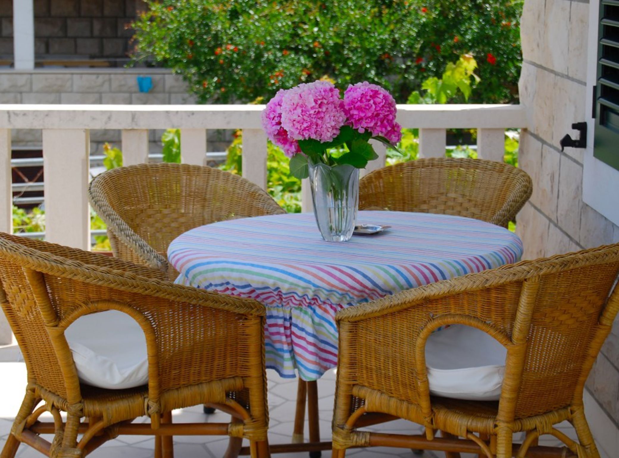 A round table with wicker chairs, a striped tablecloth, and a vase with pink flowers.