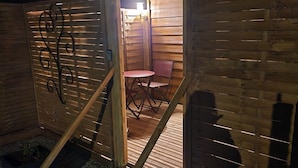 Una cabaña de madera con la puerta abierta, una mesa pequeña y una silla en su interior, y una planta en maceta en el exterior.