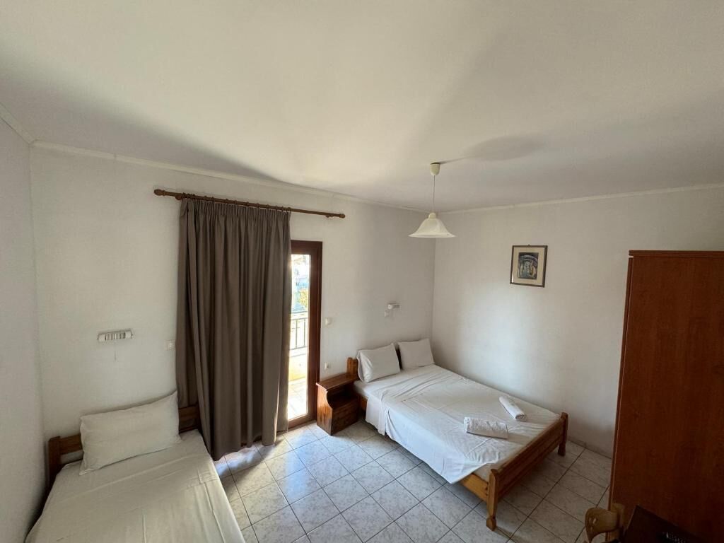Une petite chambre propre, avec deux lits, une fenêtre avec des rideaux, une table de chevet et un luminaire suspendu.