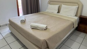 Un letto rifatto con ordine, con copriletto e cuscini color crema, un comodino in legno con un cassetto e una finestra con tende.
