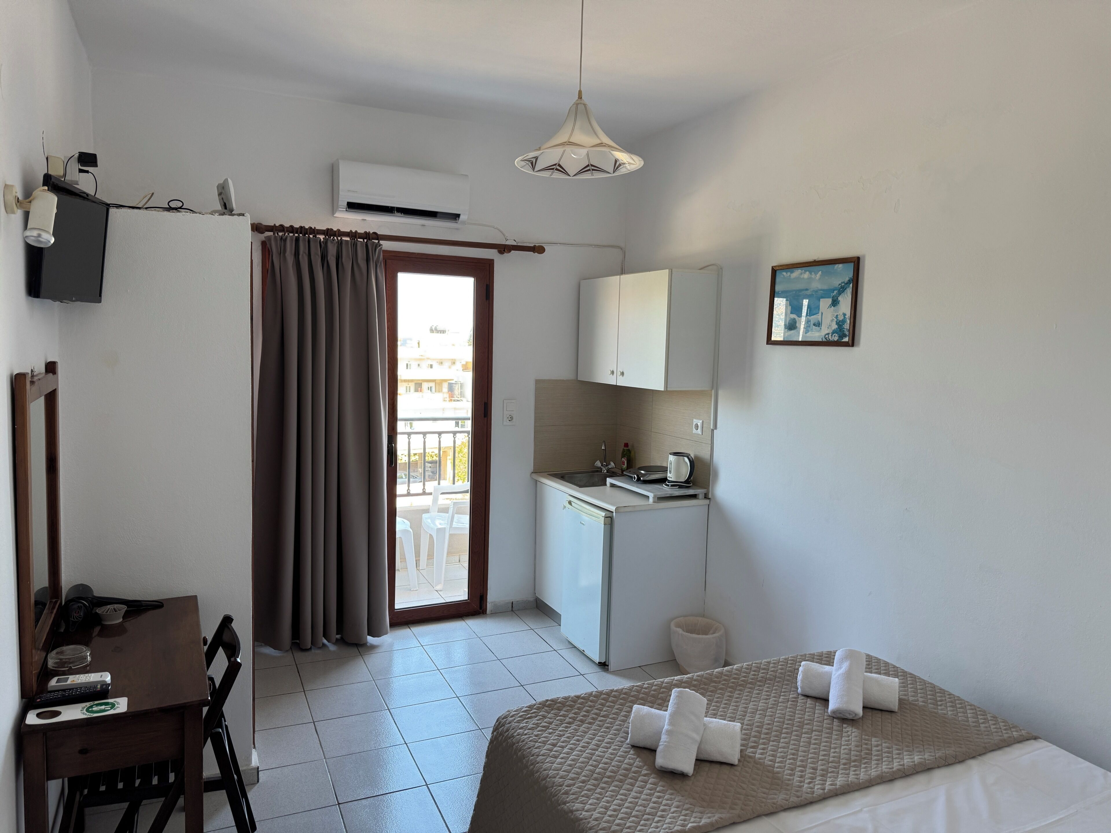 A hotel room with a bed, a desk, a chair, a kitchenette, a balcony, and a TV.