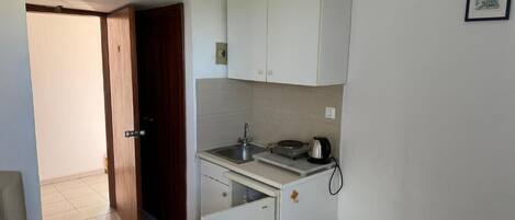 Une cuisine compacte équipée d’un évier, d’un petit réfrigérateur et d’un four à micro-ondes. Une porte donne accès à une autre pièce, et on aperçoit un lit dans un coin.