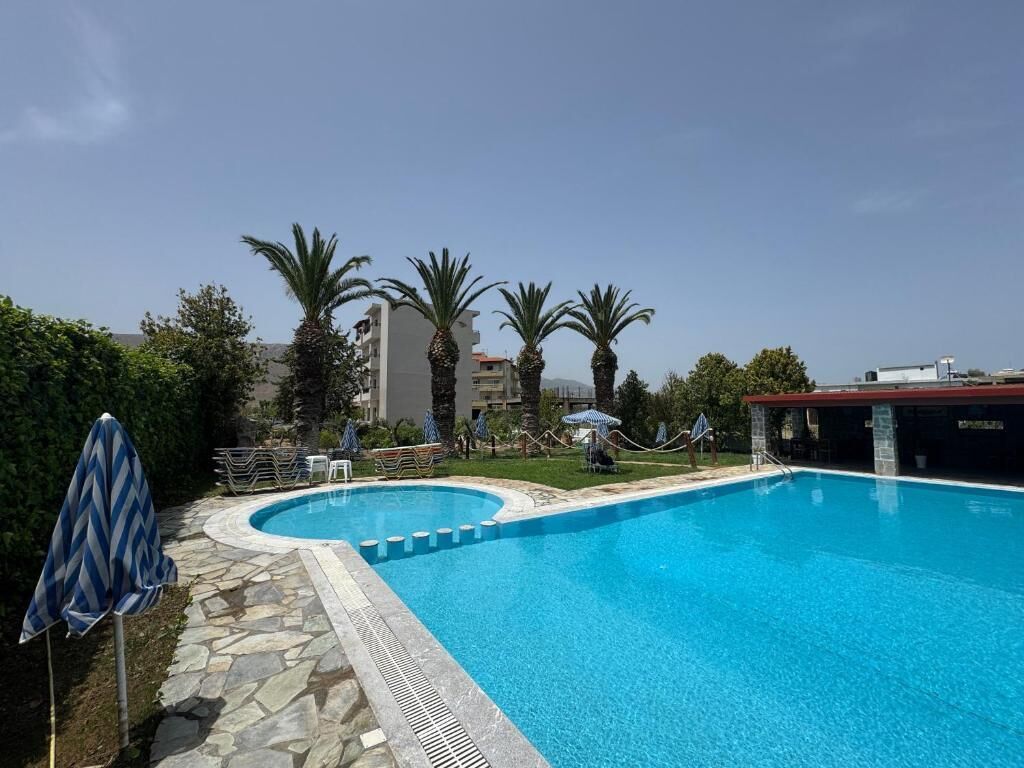 A swimming pool with stone tiles, palm trees, and a building in the background.