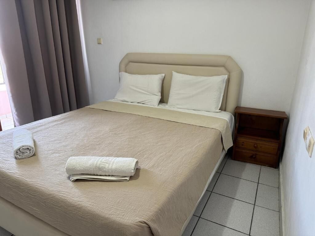 Un lit bien fait, avec une couverture beige et des oreillers, une table de chevet en bois dotée d’un tiroir, et une fenêtre avec des rideaux.