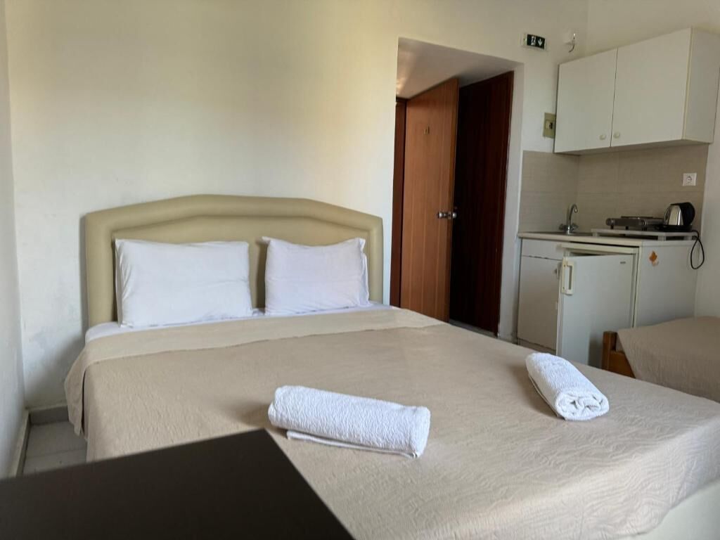 A hotel room with a bed, two towels, a wooden door, a kitchenette, and a small table.